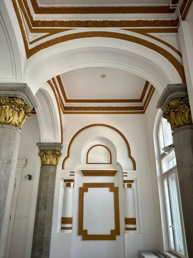 Interior view of a grand hall with white walls, gold decorative trim, and tall grey columns with gilded capitals under arches.