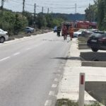 Emergency responders and a police car on a street, with a fire truck in the distance and people in red uniforms approaching a blocked road area.