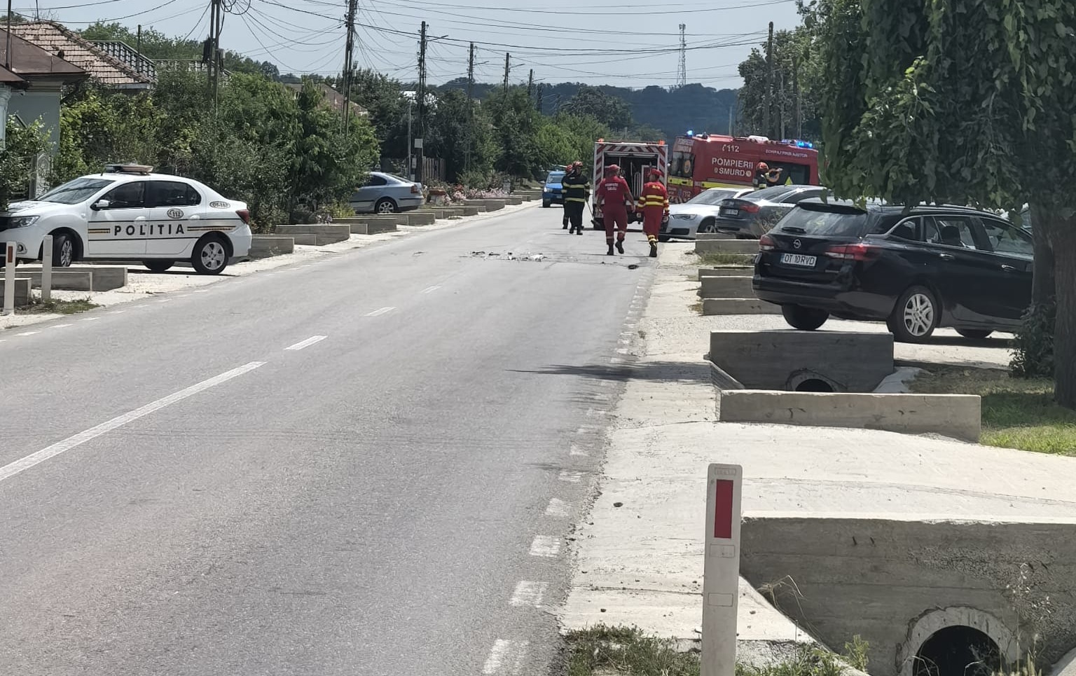 Emergency responders and a police car on a street, with a fire truck in the distance and people in red uniforms approaching a blocked road area.