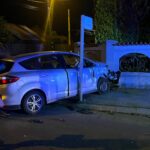 Silver sedan crashed into a low wall with severe front-end damage at night; officer standing nearby under streetlights.