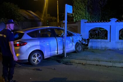 Silver sedan crashed into a low wall with severe front-end damage at night; officer standing nearby under streetlights.