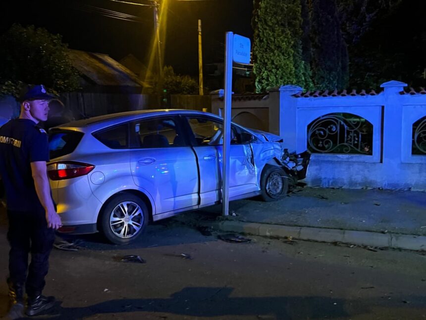 Silver sedan crashed into a low wall with severe front-end damage at night; officer standing nearby under streetlights.