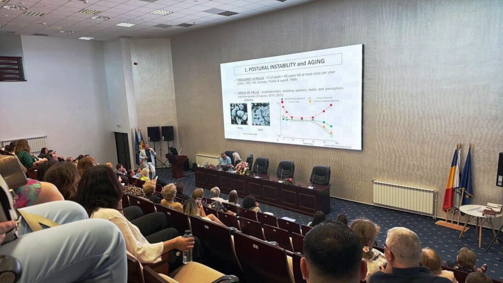 aging vertij - Jurnalul Olteniei – Știri din Craiova și Oltenia Conference lecture hall with a large screen showing a slide on postural instability and aging; audience seated and panel at front, flags on the right.