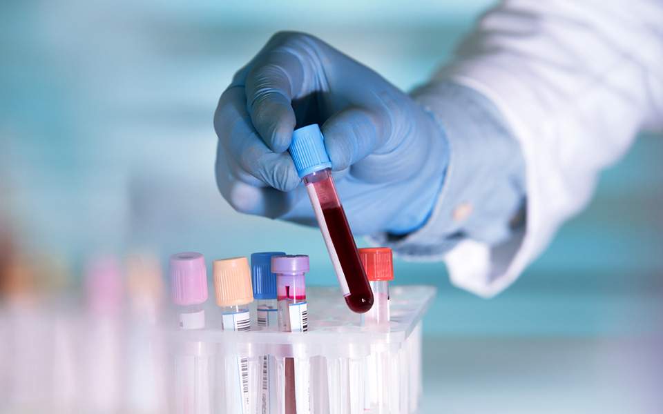 Lab technician in blue gloves places a blood sample in a test tube from a rack of vials with colored caps.