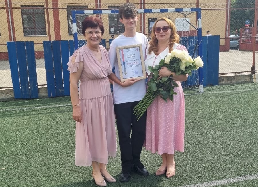 Young man in a white shirt holds a framed certificate, flanked by two women in pink dresses, one carrying a bouquet of white roses on a fenced outdoor sports field.