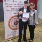 Young man in a blazer holds a diploma and orange flag while standing with a smiling woman at an academic award ceremony, with a banner in the background.