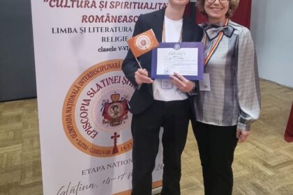 Young man in a blazer holds a diploma and orange flag while standing with a smiling woman at an academic award ceremony, with a banner in the background.