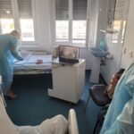 Nurse in blue gown leaning over a hospital bed papers on the bed, laptop on a small cart nearby, windowed room.
