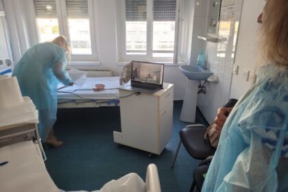Nurse in blue gown leaning over a hospital bed papers on the bed, laptop on a small cart nearby, windowed room.
