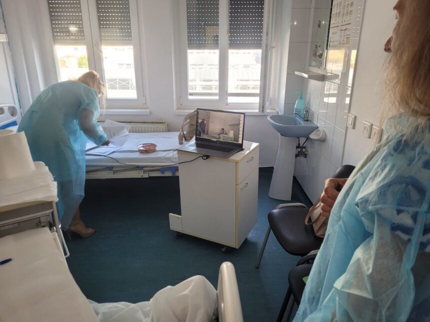 Nurse in blue gown leaning over a hospital bed papers on the bed, laptop on a small cart nearby, windowed room.