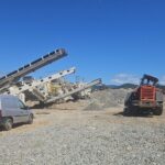 Construction site with multiple conveyor belts, a grey van, and an orange excavator under a clear blue sky.
