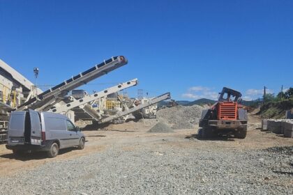 Construction site with multiple conveyor belts, a grey van, and an orange excavator under a clear blue sky.