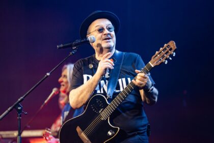 Male guitarist wearing a black hat and sunglasses performs on stage with a black electric guitar and microphone nearby.