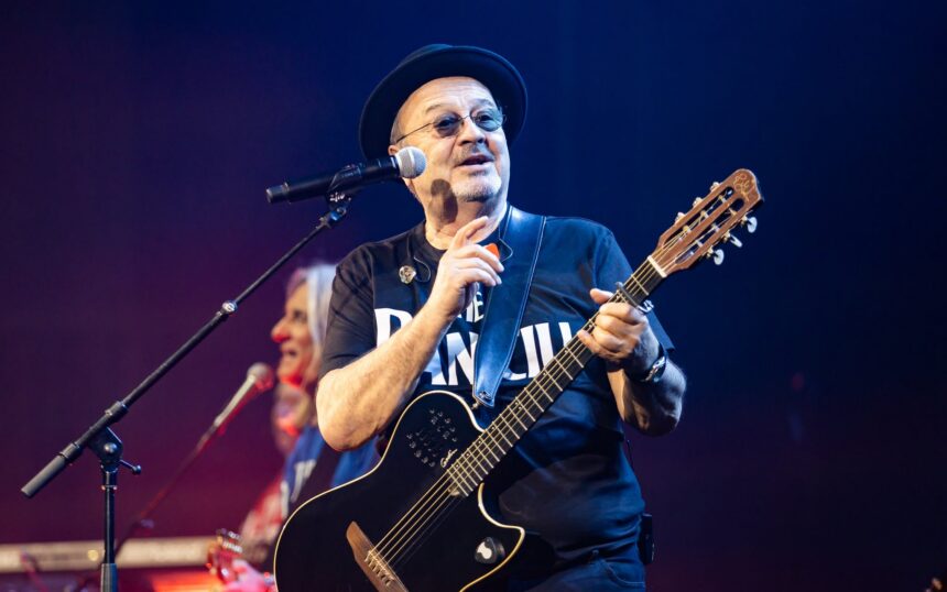 Male guitarist wearing a black hat and sunglasses performs on stage with a black electric guitar and microphone nearby.