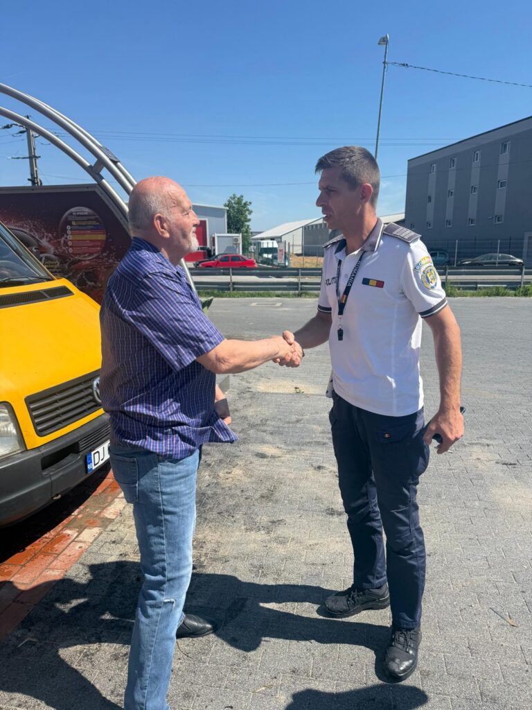 Two men shake hands in a parking lot beside a yellow van; one is a civilian in a blue checkered shirt, the other a uniformed officer.