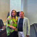 Two adults pose for a photo in a conference room; the woman wears a bright green tropical blouse and stands beside a man in a striped blazer by a flower-adorned desk.