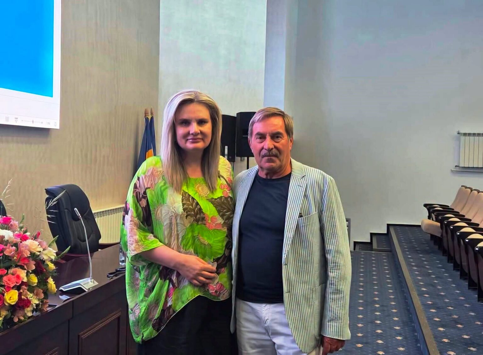 Two adults pose for a photo in a conference room; the woman wears a bright green tropical blouse and stands beside a man in a striped blazer by a flower-adorned desk.