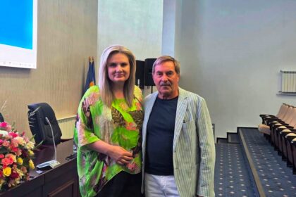 Two adults pose for a photo in a conference room; the woman wears a bright green tropical blouse and stands beside a man in a striped blazer by a flower-adorned desk.