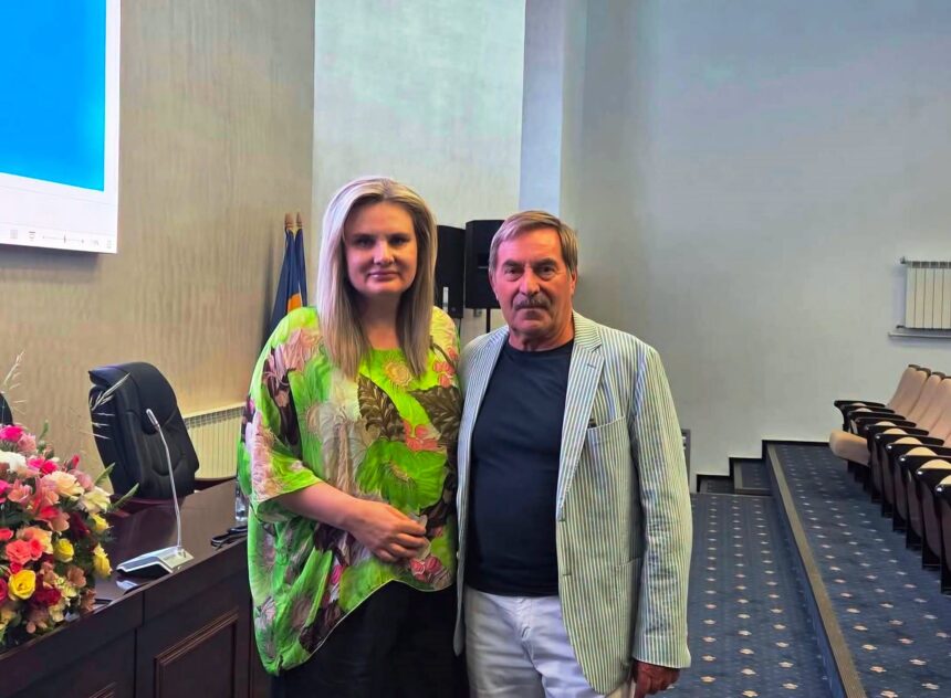 Two adults pose for a photo in a conference room; the woman wears a bright green tropical blouse and stands beside a man in a striped blazer by a flower-adorned desk.