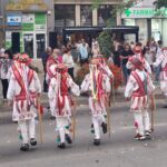 Dancers in white outfits with red embroidery and colorful ribbons march down a street while spectators watch.