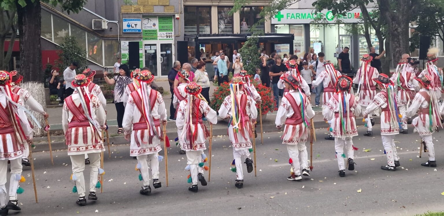 Dancers in white outfits with red embroidery and colorful ribbons march down a street while spectators watch.