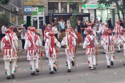 Dancers in white outfits with red embroidery and colorful ribbons march down a street while spectators watch.