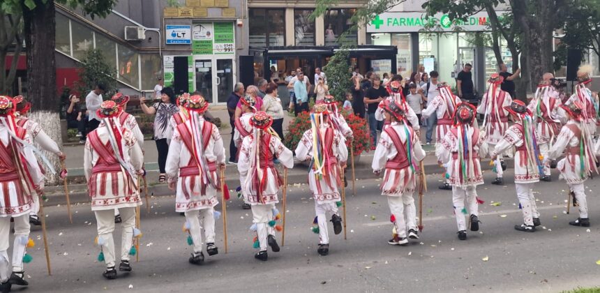 Dancers in white outfits with red embroidery and colorful ribbons march down a street while spectators watch.