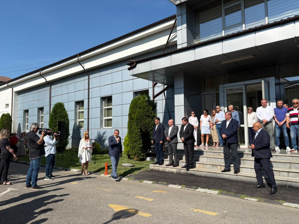 Group of people in business attire standing on the steps outside a modern building for a ceremony; journalists with cameras on the left.