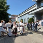 Group of dancers in traditional white folk costumes performing outside a modern building, while spectators watch on the steps.