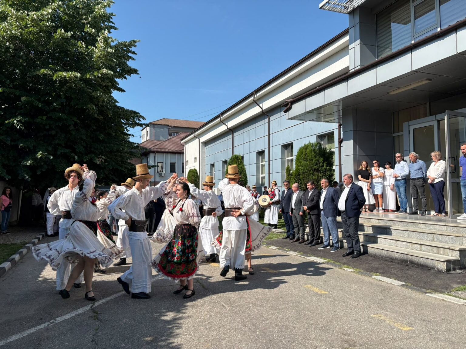 Group of dancers in traditional white folk costumes performing outside a modern building, while spectators watch on the steps.