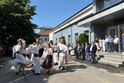 Group of dancers in traditional white folk costumes performing outside a modern building, while spectators watch on the steps.