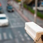Close-up of a white street camera mounted on a pole overlooking a busy city street with cars and crosswalks in soft focus behind it.