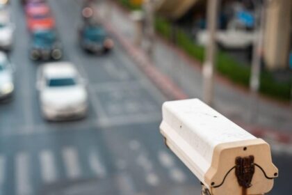 Close-up of a white street camera mounted on a pole overlooking a busy city street with cars and crosswalks in soft focus behind it.