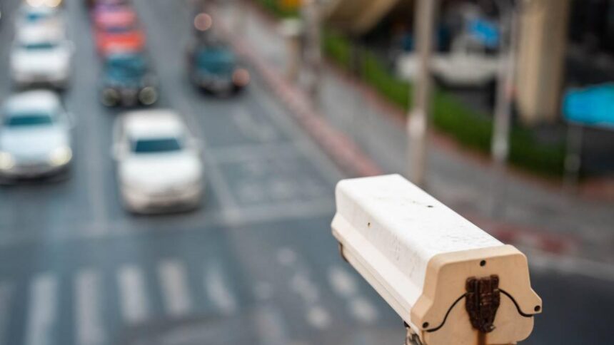 Close-up of a white street camera mounted on a pole overlooking a busy city street with cars and crosswalks in soft focus behind it.