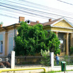 Old yellow building with ornate façade partially obscured by trees, iron fence in foreground and overhead power lines.