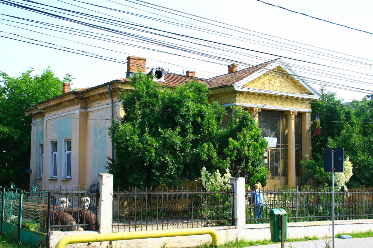 Old yellow building with ornate façade partially obscured by trees, iron fence in foreground and overhead power lines.