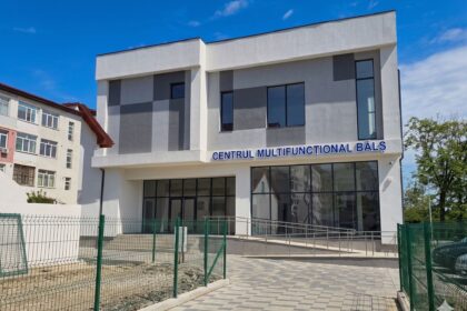 Modern gray-and-white building with large glass windows and a blue sign reading CENTRUL MULTIFUNCTIONAL BALS under a clear blue sky.