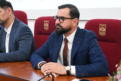 Bearded man in a blue checkered suit, white shirt, red tie and glasses seated at a conference table with a microphone in front.