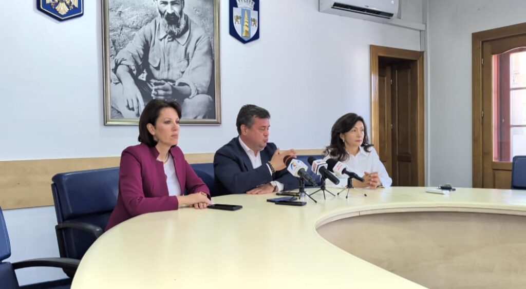 Three officials seated at a curved conference table giving a press briefing; microphones are set up in front of them, a portrait and shields on the wall behind.