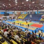 Indoor gymnasium with multicolored mats for a martial arts tournament; spectators seated in yellow and blue stands surround the arena.