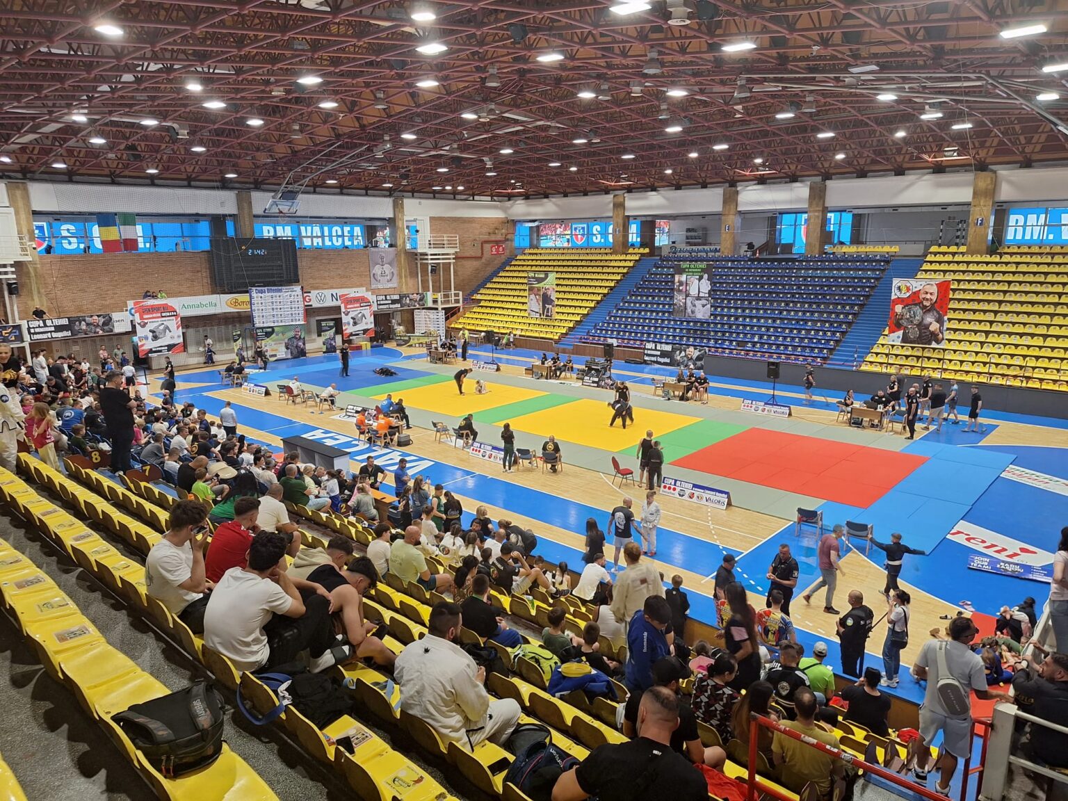 Indoor gymnasium with multicolored mats for a martial arts tournament; spectators seated in yellow and blue stands surround the arena.