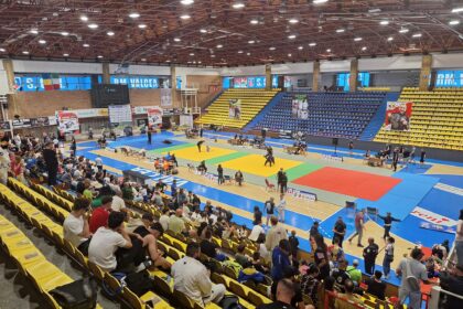 Indoor gymnasium with multicolored mats for a martial arts tournament; spectators seated in yellow and blue stands surround the arena.