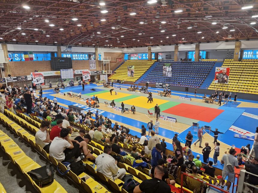 Indoor gymnasium with multicolored mats for a martial arts tournament; spectators seated in yellow and blue stands surround the arena.