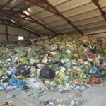 Large pile of mixed trash and garbage bags piled inside an industrial warehouse with metal support beams above.