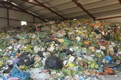 Large pile of mixed trash and garbage bags piled inside an industrial warehouse with metal support beams above.