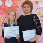 Girl and woman stand together, each holding diplomas, in front of a pink floral backdrop with heart shapes.