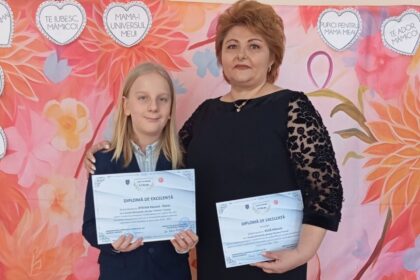 Girl and woman stand together, each holding diplomas, in front of a pink floral backdrop with heart shapes.