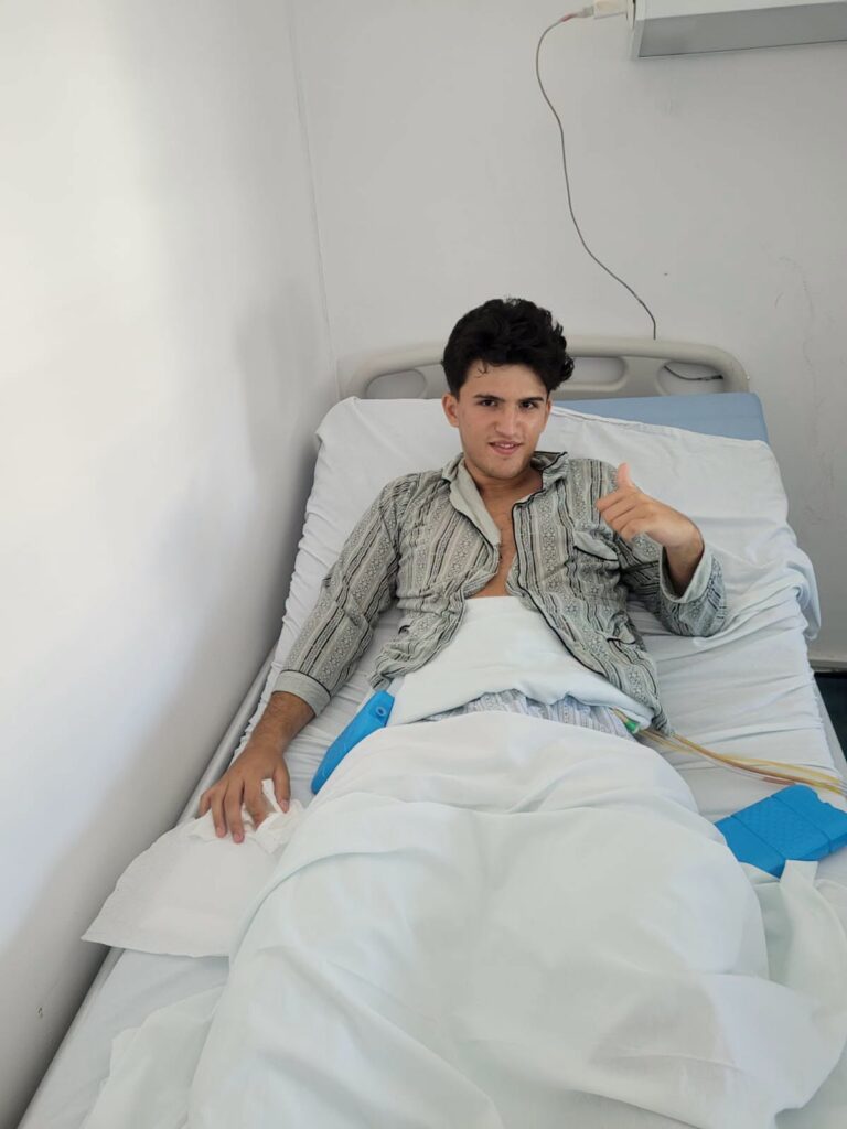Young man in a hospital bed sits up under white linens, giving a thumbs-up with medical tubing nearby to indicate recovery.