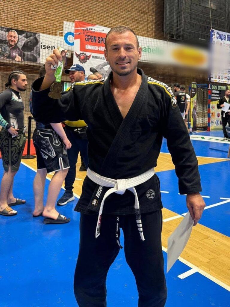 Martial artist in a black gi and white belt smiles while holding up a gold medal in a gym during a competition, with others and banners in the background.