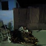 Man kneeling on a patterned blanket at night near a wooden gate, with shoes and a chair nearby in a dim outdoor area.